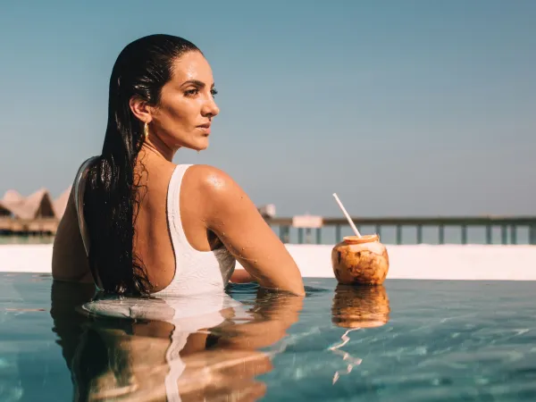 Woman leaning on pool edge with coconut drink, serenely gazing at overwater jetty at JOALI Maldives.