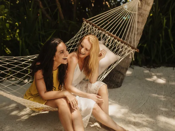 Two women laughing in a hammock under palm trees on a sunny JOALI Maldives beach