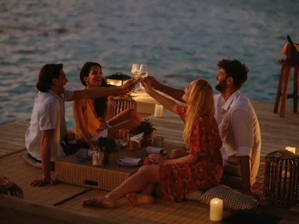 Group of friends raising wine glasses on a wooden jetty over lagoon waters at sunset at JOALI Maldives.